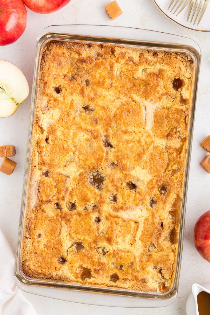 Easy caramel apple dump cake in casserole dish sitting on counter surrounded by apples and caramels.  