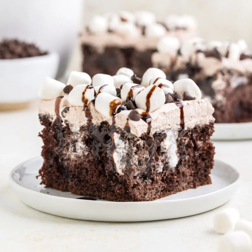 Slice of cake on a plate in the foreground with two slices of cake and a mug of hot chocolate in the background.