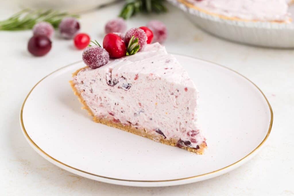 Slice of no-bake cranberry pie with sugared cranberries on plate; whole pie in background.