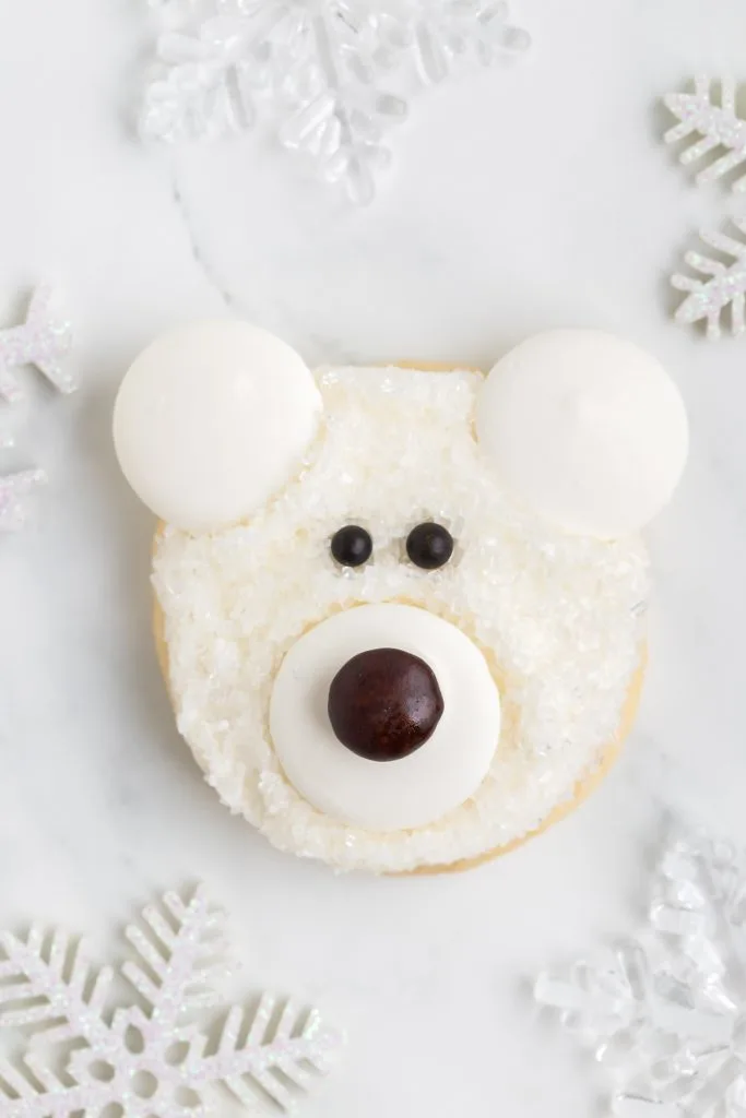 A single polar bear face cookie with a brown nose laying flat on the counter and surrounded by snow flake decorations.