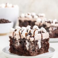 Slice of cake on a plate in the foreground with two slices of cake and a mug of hot chocolate in the background.