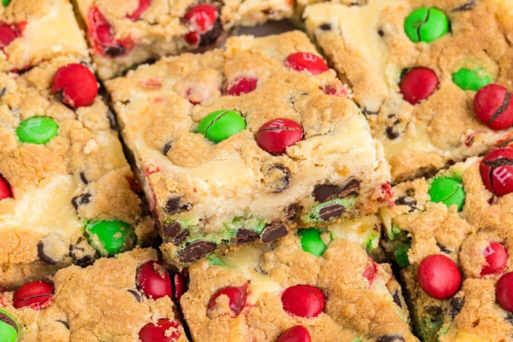 Close-up of cookie cheesecake bars topped with red, green candy-coated chocolates and chocolate chips, cut into squares.