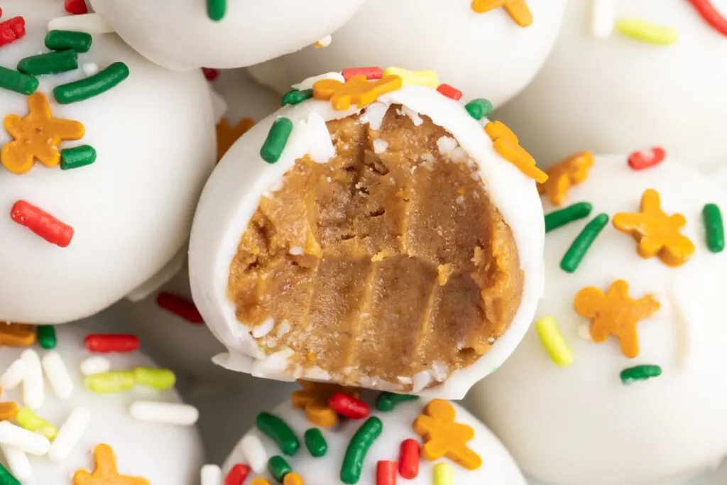 Close-up of white chocolate-covered gingerbread truffles, one is bitten to reveal a peanut butter center. Decorated with colorful sprinkles, including red, yellow, and green shapes, the truffles are arranged closely together.