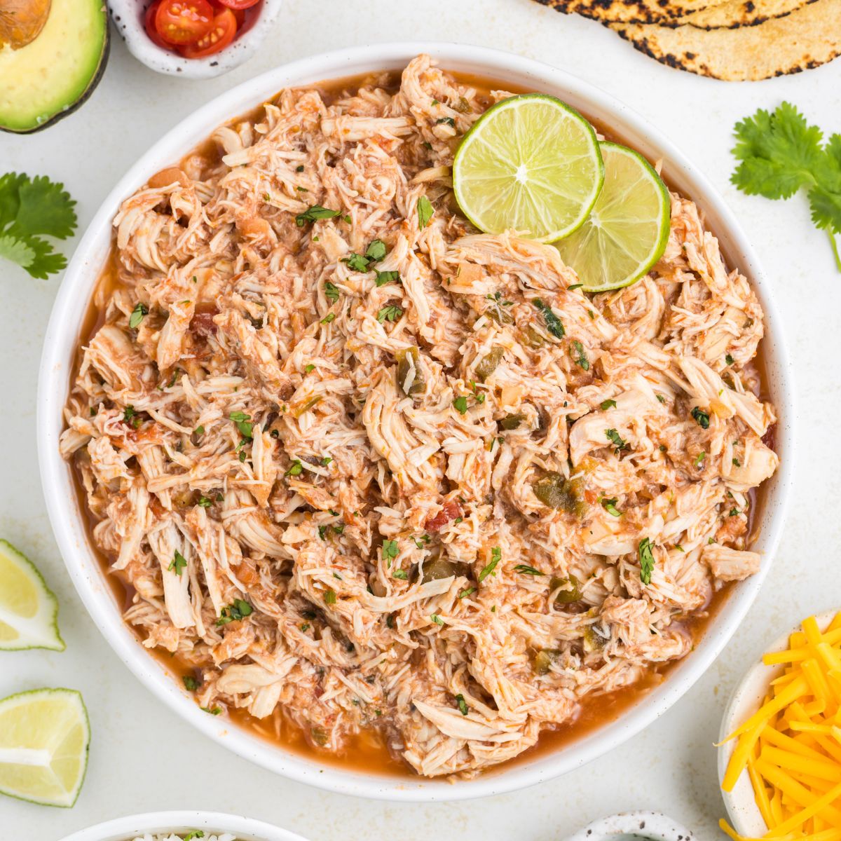 Bowl of slow cooker salsa chicken surrounded by tortillas, rice and condiments.