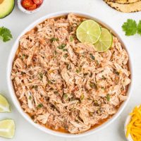 Bowl of slow cooker salsa chicken surrounded by tortillas, rice and condiments.