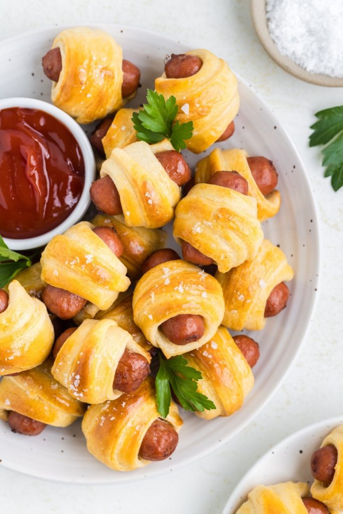 Pigs in a blanket stacked on a plate with a ramekin of ketchup. 