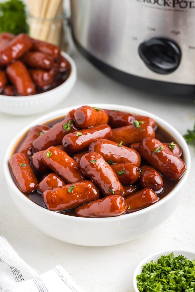 Bowl of little smokies cooked with grape jelly on the counter with slow cooker in the background. 