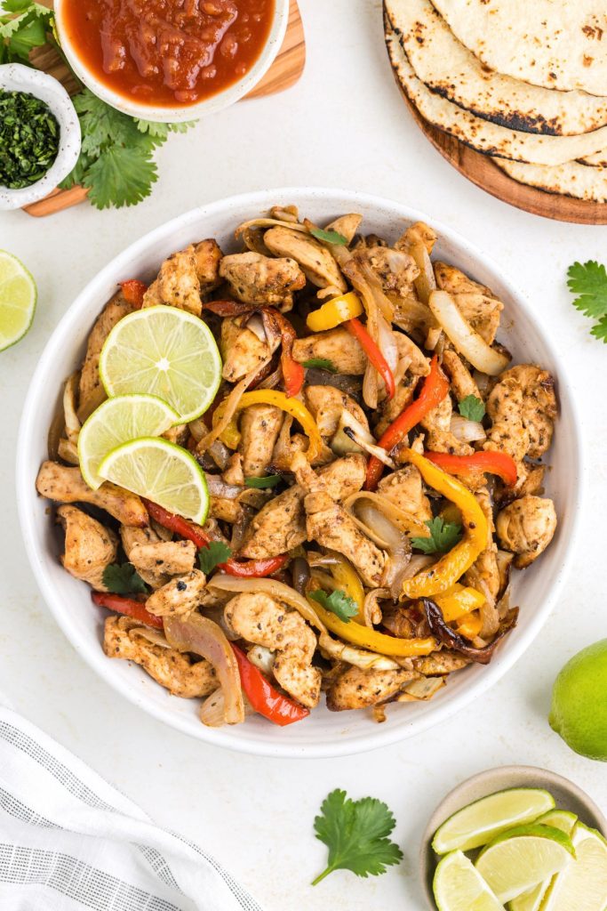 Air fryer chicken fajitas in a bowl on the counter surrounded by tortillas, salsa and lime.