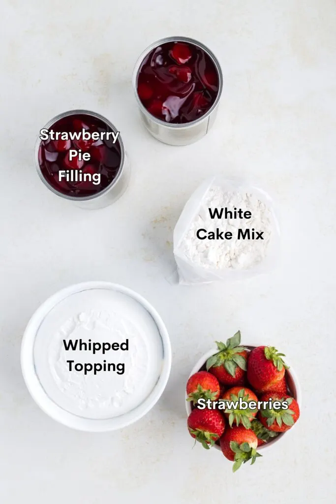 Ingredients such as white cake mix, strawberry pie filling, whipped topping, and fresh strawberries displayed on the counter.