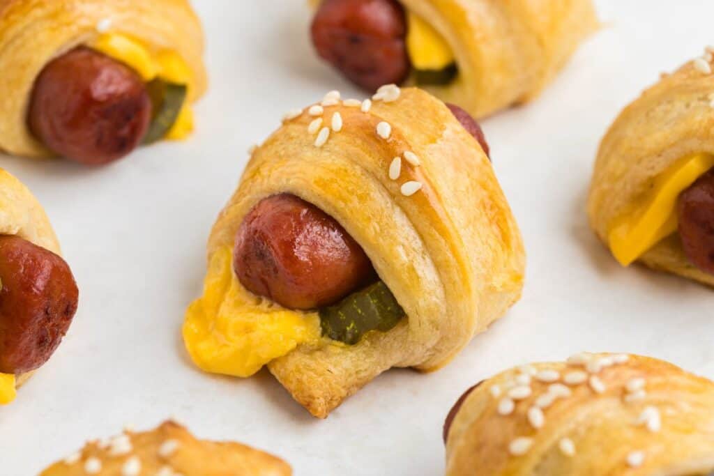 Crescent roll cheeseburger pigs in a blanket with cheese, pickles, and sesame seeds on a white surface.