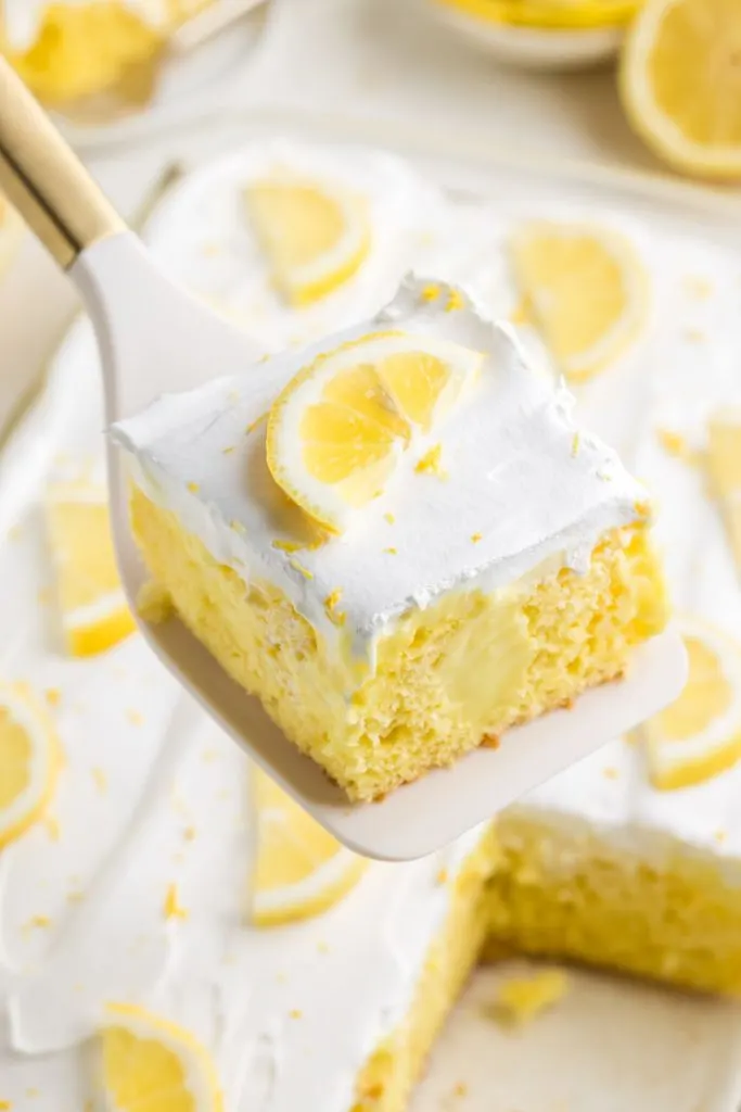 Slice of lemon pudding poke cake topped with a piece of lemon being removed from the pan. 