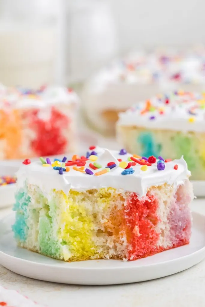Slice of vibrant rainbow poke cake with a forkful missing.