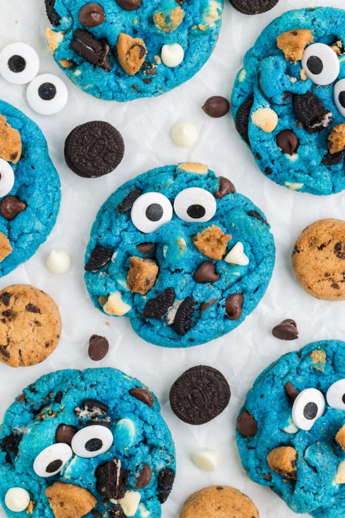 Cookie Monster cookies displayed on the counter surrounded by mini chocolate chip cookies, Oreos and white and chocolate chips.
