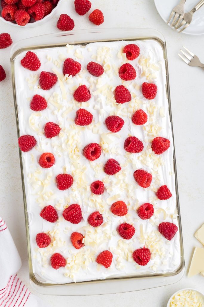 Raspberry white chocolate poke cake in a pan covered in fresh raspberries and white chocolate curls.