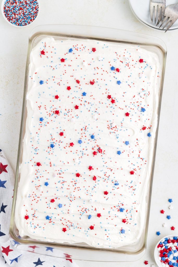 Red, white and blue poke cake in a cake pan with white chocolate frosting and sprinkles.