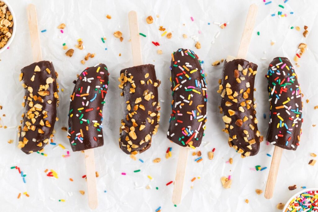 Chocolate banana popsicles with sprinkles and nuts arranged in a row.