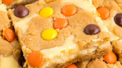 Peanut butter cheesecake cookie bars topped with orange, yellow, and brown Reese's Pieces chocolates; one bar is lifted.