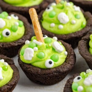 Chocolate Halloween cauldron cookie cups with green frosting, candy eyes, skulls, pearls, and pretzel sticks on a light surface.