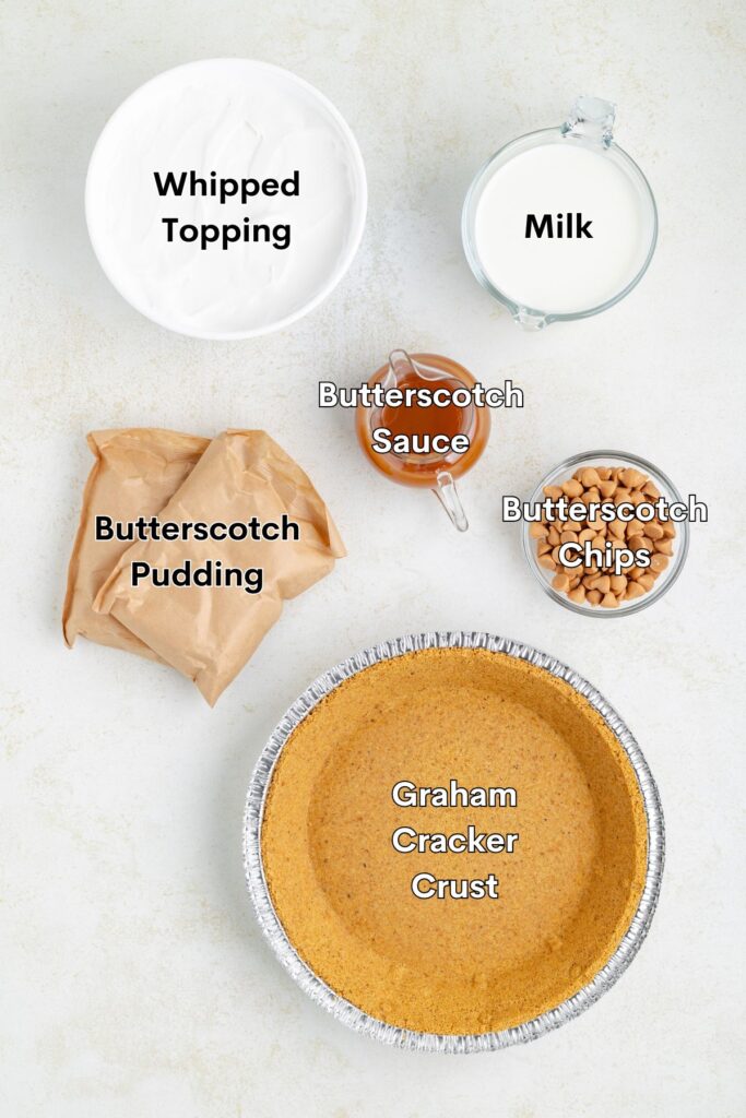 Ingredients such as graham cracker pie crust, butterscotch pudding, milk, whipped topping, butterscotch sauce and butterscotch chips arranged on the counter.