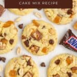 Snickers Cake Mix Cookies with Snickers chunks, chocolate chips, caramel, and peanuts on a white surface with a Snickers bar.