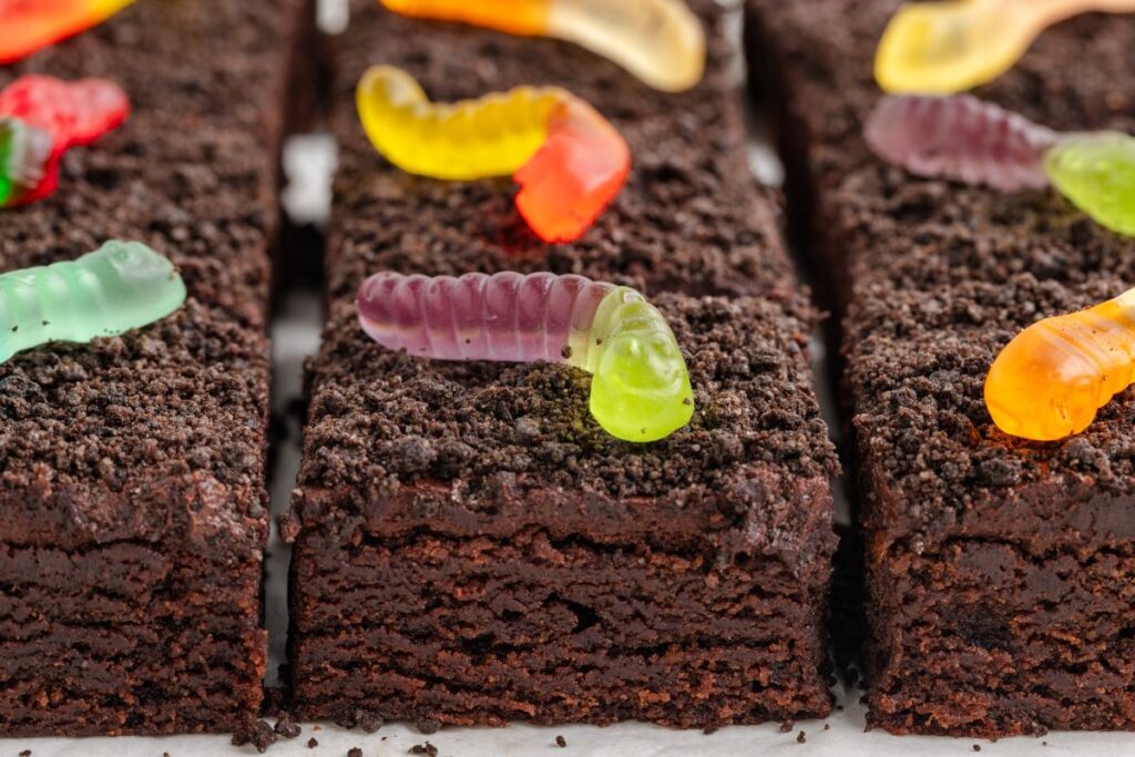 Rows of brownies with chocolate frostings, crushed Oreos and candy worms.