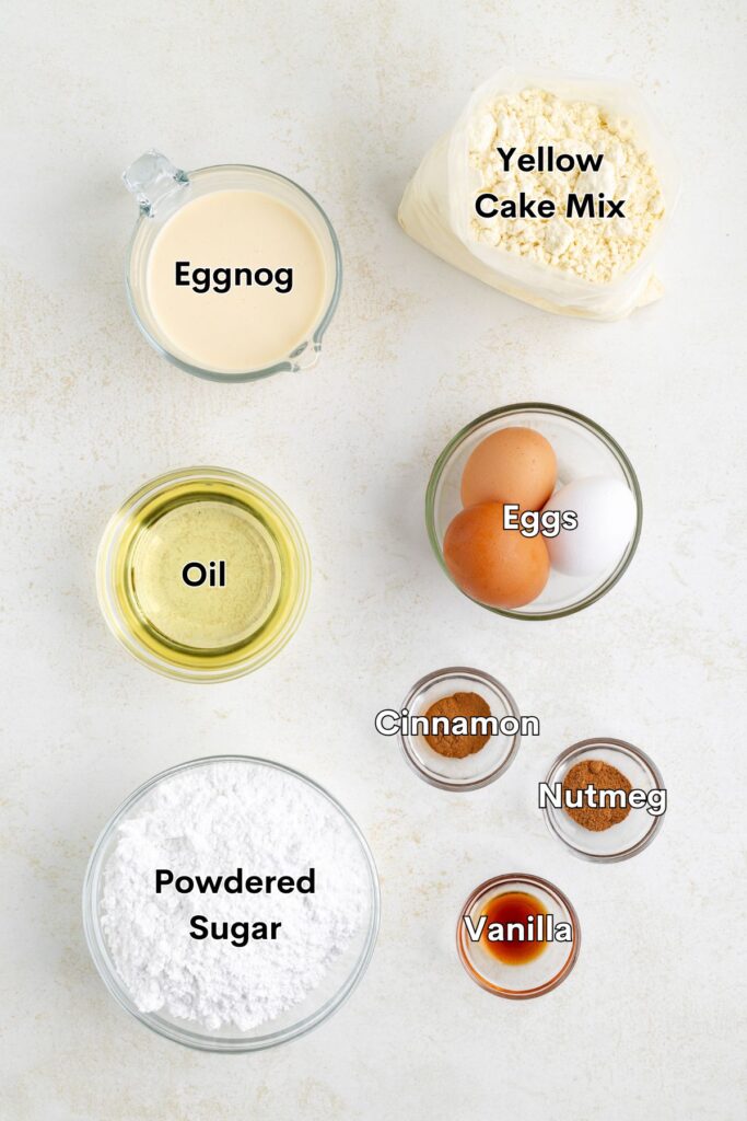 Ingredients such as yellow cake mix, eggnog, oil, eggs, vanilla, gound nutmeg, ground cinnamon, and powdered sugar arranged in bowls.