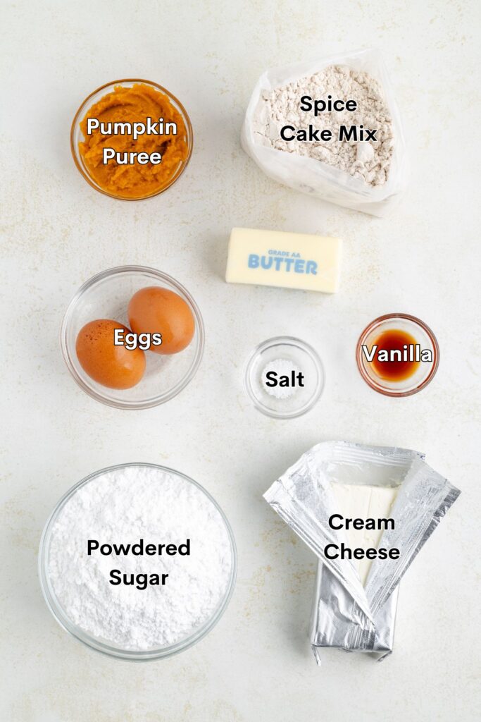 Ingredients such as spice cake mix, pumpkin puree, eggs, butter, cream cheese, powdered sugar, vanilla, and salt arranged on the counter.