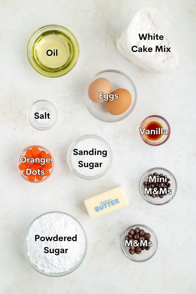 Ingredients such as white cake mix, eggs, oil, powdered sugar, butter, vanilla, salt, sanding sugar, Dot candies, and M&Ms displayed on the counter.