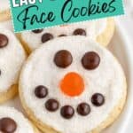 Snowman face cookies with white frosting, chocolate eyes and mouths, and orange noses on a white plate.
