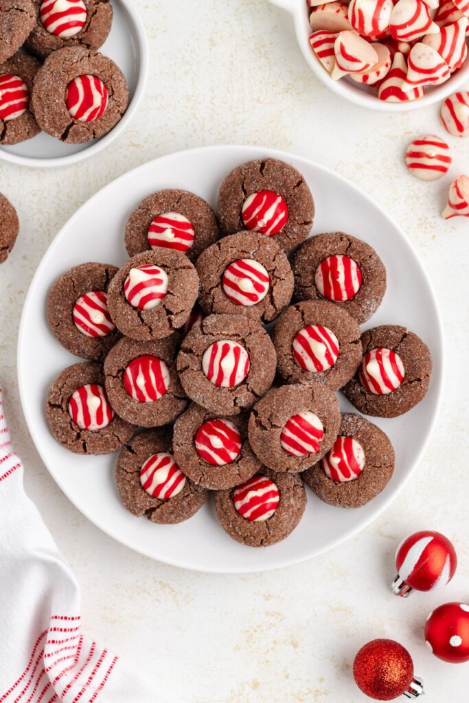 A plate of chocolate cookies topped with red and white striped candy pieces. Next to the plate, there is a bowl containing more candies. A red and white striped cloth and small red ornaments are also visible.