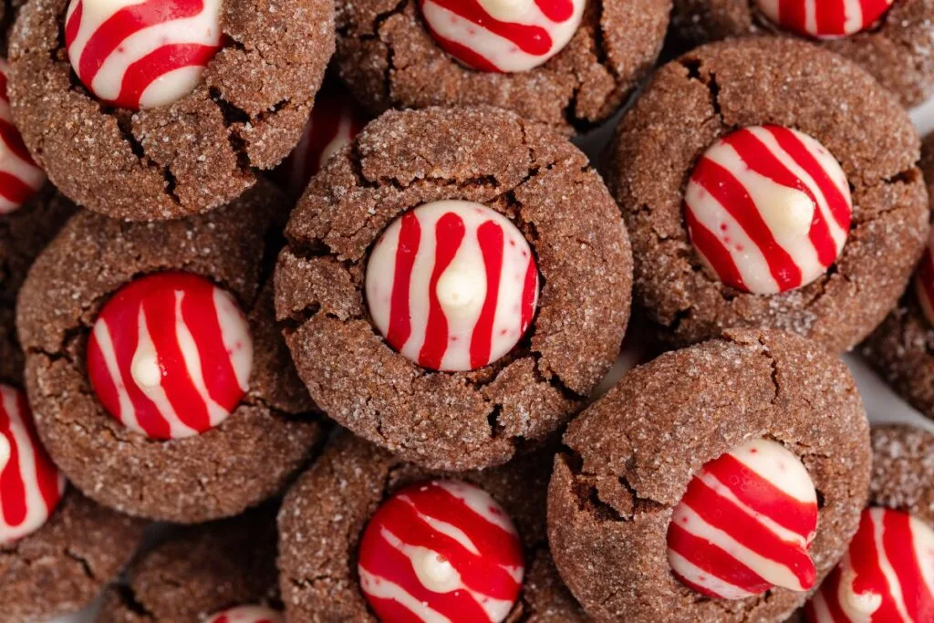 Close-up of Chocolate Peppermint Kiss Cookies, topped with white and red striped candy cane kisses. The cookies are covered in a layer of sugar, creating a festive appearance. They are arranged in a pile, highlighting their decorative toppings.