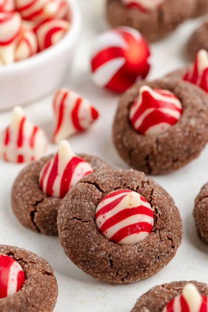 Close-up of Chocolate Peppermint Kiss Cookies topped with red and white striped chocolate kisses. A bowl filled with the candies is blurred in the background. The cookies are arranged on a light surface, showcasing their festive decoration.