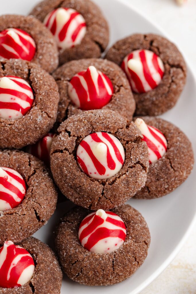 A plate of chocolate thumbprint cookies topped with white chocolate candies decorated with red swirls. The cookies are arranged closely together, showcasing their festive and colorful design.