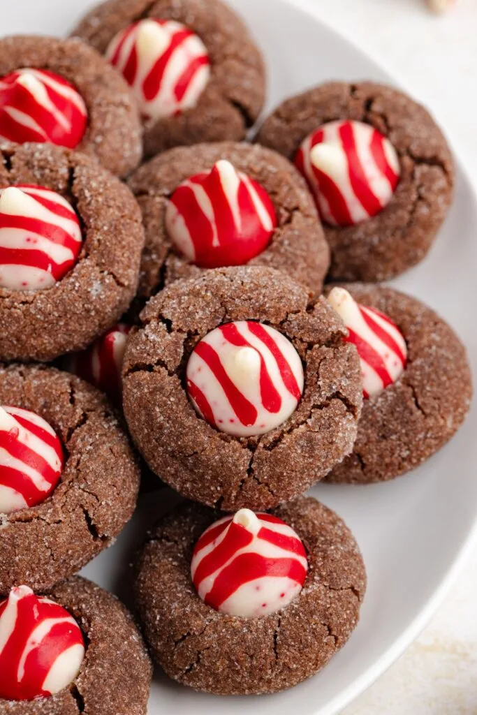 A plate of chocolate thumbprint cookies topped with white chocolate candies decorated with red swirls. The cookies are arranged closely together, showcasing their festive and colorful design.