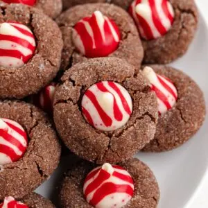 A plate of Chocolate Peppermint Kiss Cookies topped with red and white striped chocolate candies. The cookies are round and have a crackled texture, with the peppermint-flavored candies perfectly perched at the center of each delightful cookie.