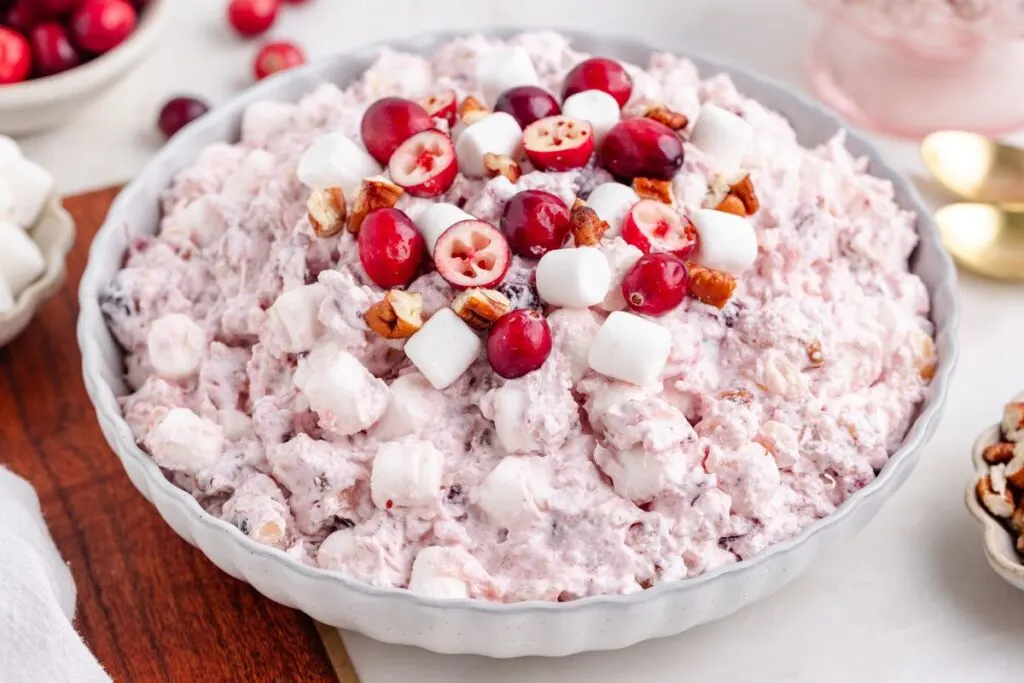 A white dish filled with cranberry fluff salad showcases a pink creamy mixture adorned with fresh cranberries, mini marshmallows, and pecan pieces. Nearby, whole cranberries accompany the scene alongside a couple of golden spoons on a rustic wooden surface.