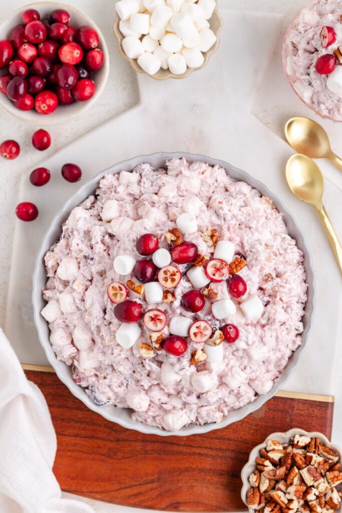 A bowl of fluffy pink salad topped with halved cranberries, mini marshmallows, and pecans. Surrounded by small bowls of whole cranberries, marshmallows, and pecans. Two gold spoons are placed nearby on a marble surface.