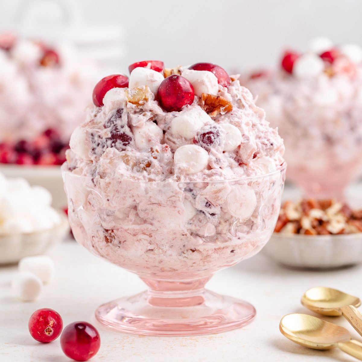 A glass dessert dish filled with a cranberry fluff salad, topped with cranberries and mini marshmallows. There are bowls of marshmallows and chopped nuts in the background, along with two gold spoons on the table.