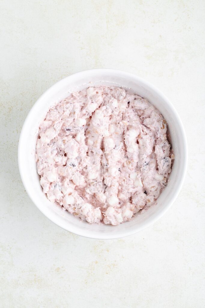 A white ceramic bowl filled with a creamy, textured mixture of fruit salad. The salad appears to contain small pieces of fruit and marshmallows, creating a pinkish hue against the light background.