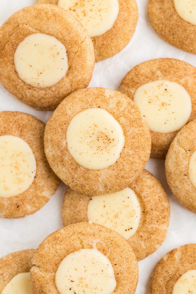 A close-up of several eggnog thumbprint cookies with a creamy white center, dusted with a sprinkle of spices. The cookies are arranged on a light surface, showcasing their round shape and textured appearance.