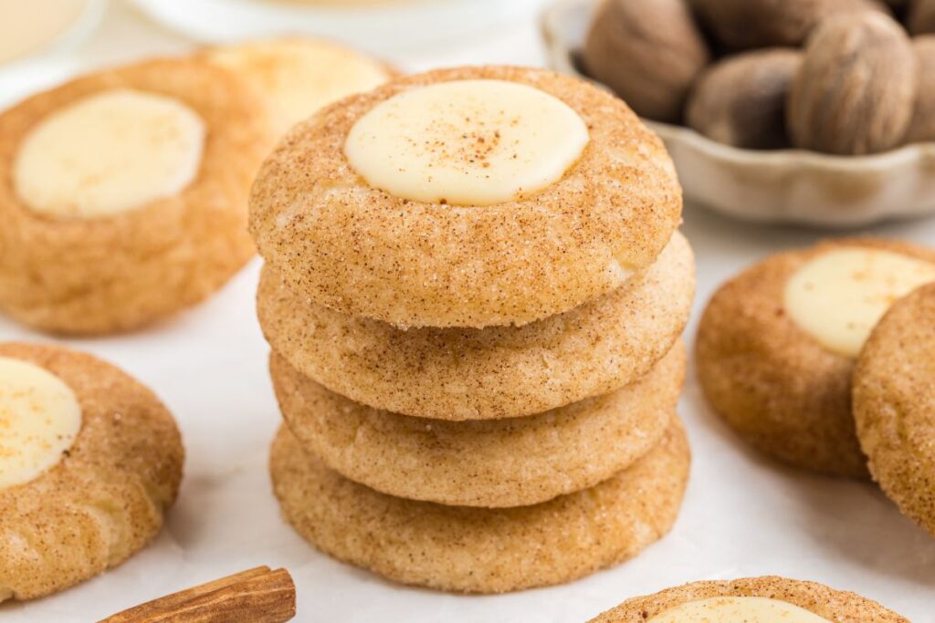 A stack of eggnog thumbprint cookies is surrounded by more cookies and a bowl of whole nutmeg. The cookies are sprinkled with cinnamon and have an eggnog center. A cinnamon stick rests nearby.