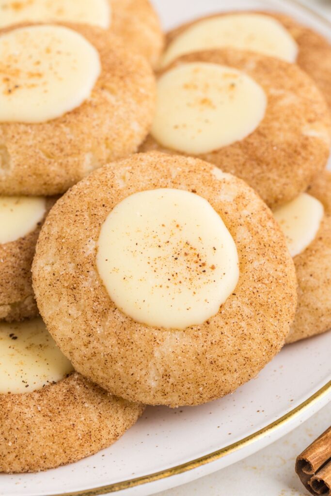 A plate of eggnog thumbprint cookies with a creamy eggnog center, dusted with cinnamon sugar, arranged closely together. The cookies have a soft, round shape with a golden-brown hue.