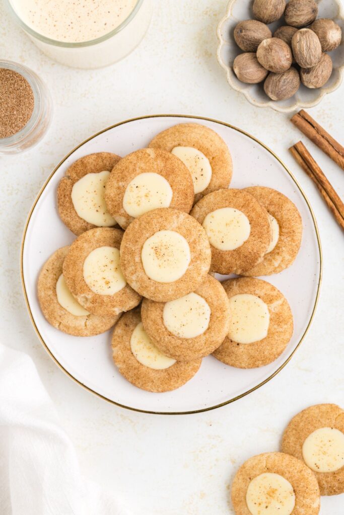 A plate of thumbprint snickerdoodle cookies with creamy eggnog ganache and a sprinkle of nutmeg is surrounded by a cup of eggnog, whole nutmegs in a bowl, and cinnamon sticks on a light surface.