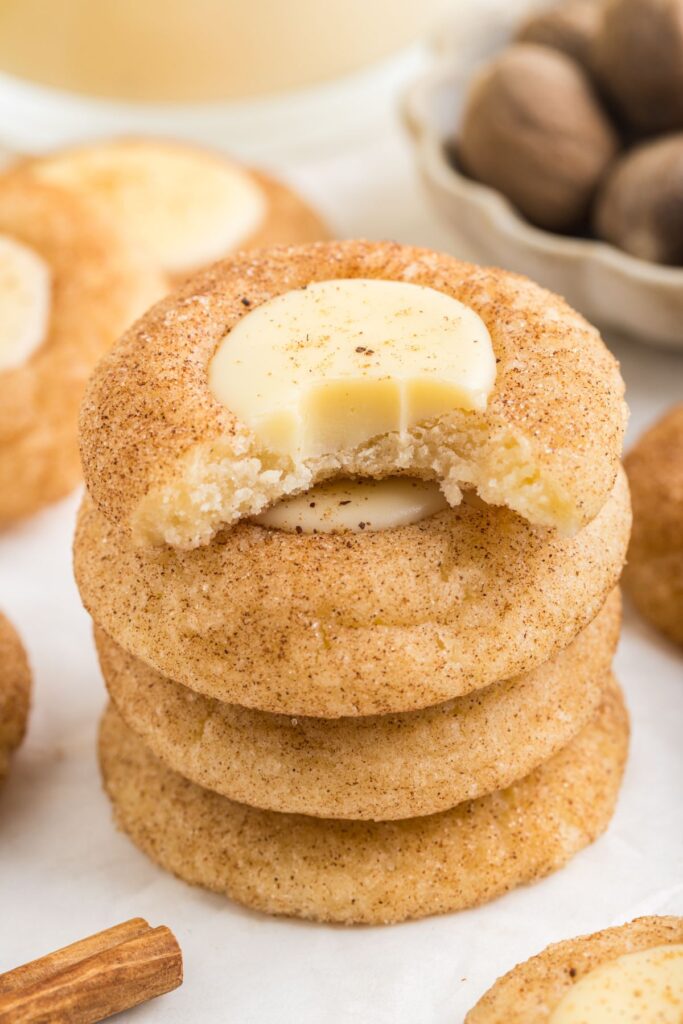 A stack of eggnog thumbprint cookies topped with eggnog white chocolate ganache. The top cookie has a bite taken out, revealing a soft texture. In the background, a bowl of whole nutmegs and a cinnamon stick are visible.