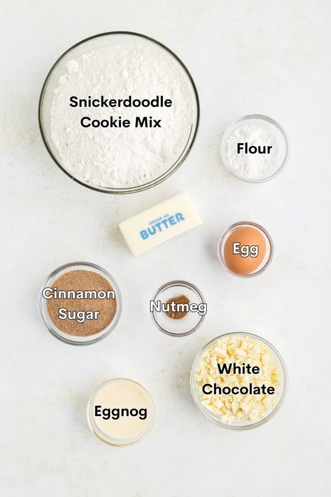 Top view of baking ingredients including labeled bowls of snickerdoodle cookie mix, flour, cinnamon sugar, nutmeg, eggnog, and white chocolate. A stick of butter and an egg are also displayed on a light surface.