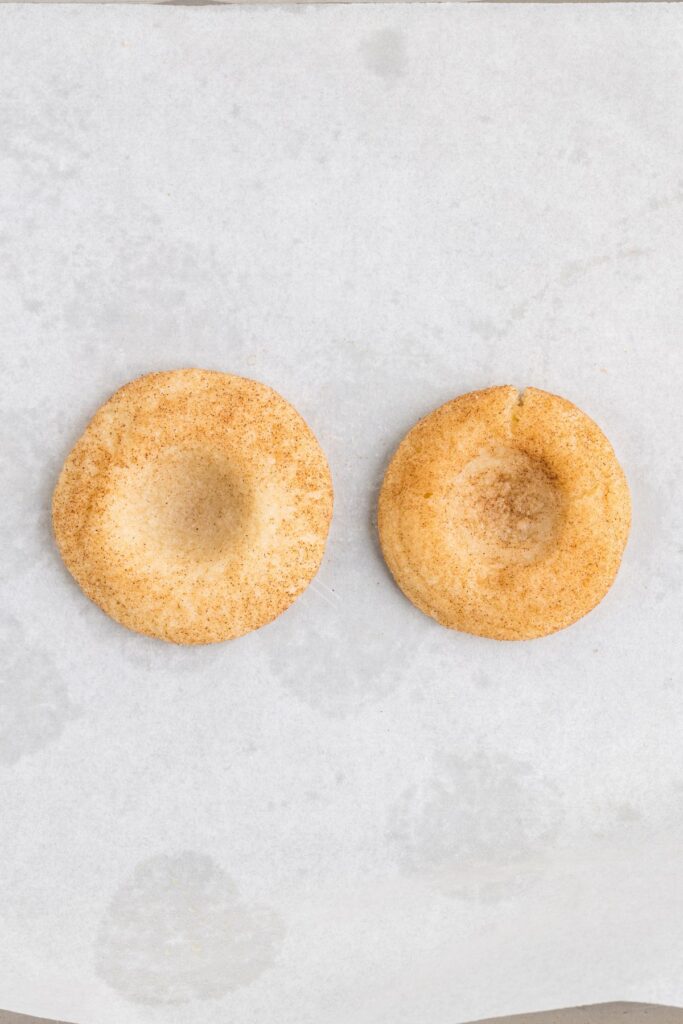 Two round cookies are placed on white parchment paper. Both cookies are lightly dusted with cinnamon sugar, giving them a slightly brown, speckled appearance. They have indented centers, suggesting they may be thumbprint cookies.