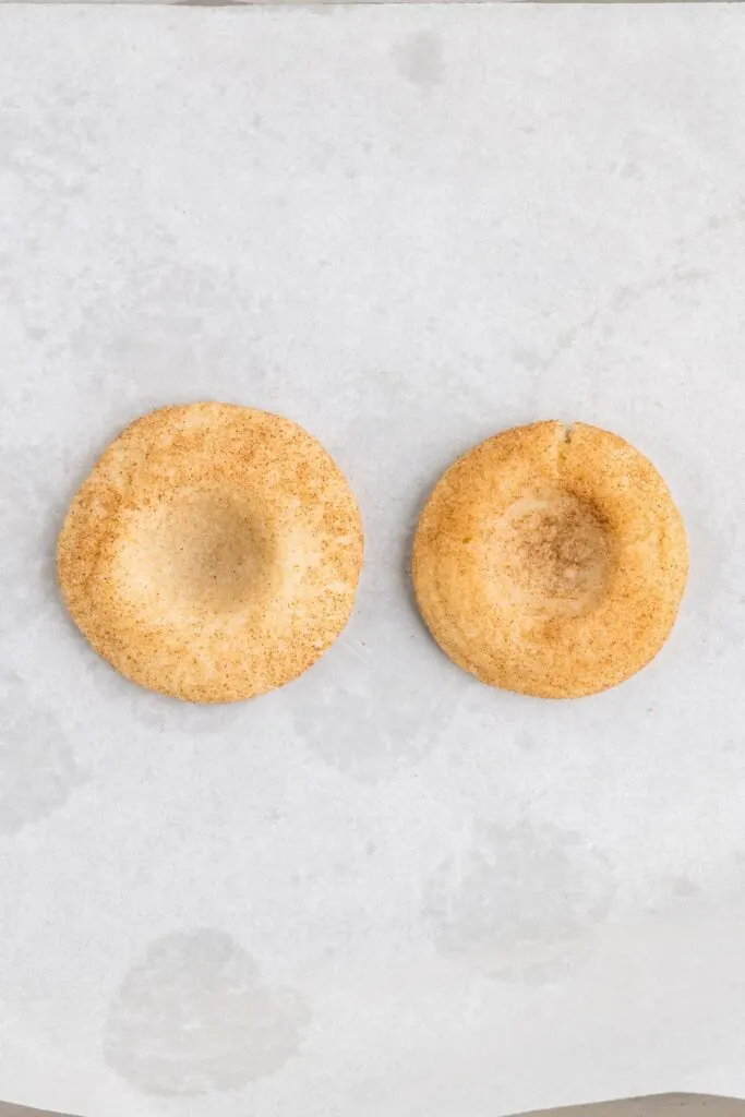 Two round cookies are placed on white parchment paper. Both cookies are lightly dusted with cinnamon sugar, giving them a slightly brown, speckled appearance. They have indented centers, suggesting they may be thumbprint cookies.