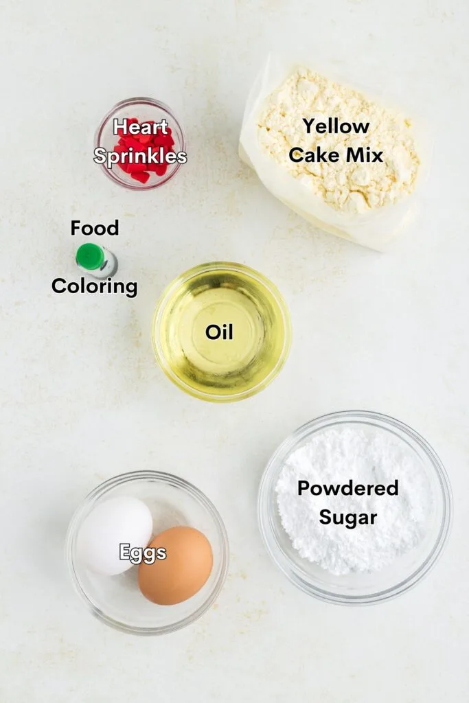 Yellow cake mix, eggs, oil, powdered sugar, green food coloring, and red heart sprinkles arranged on the counter.