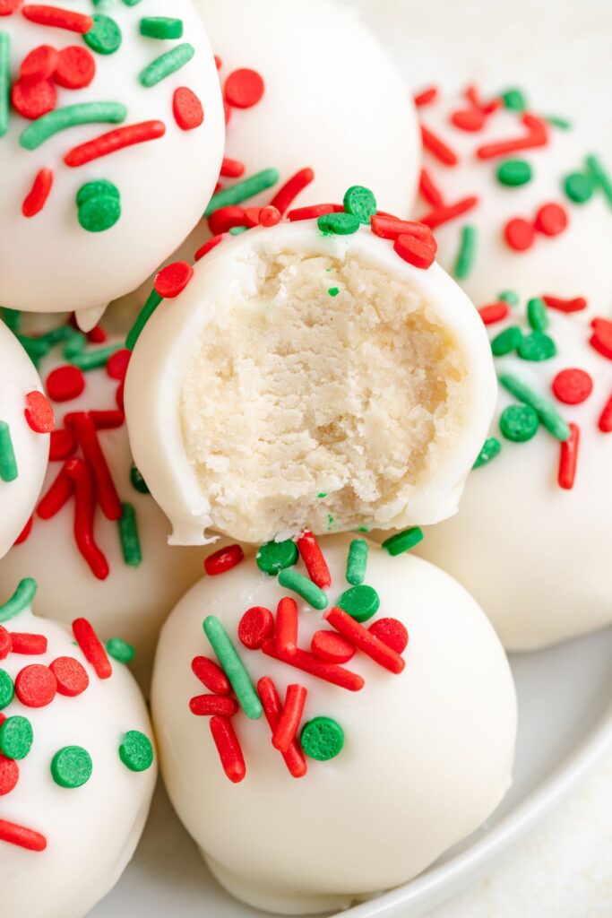 Sugar cookie truffles truffles topped with red and green sprinkles are stacked on a plate. One truffle is cut open, showing a creamy interior.