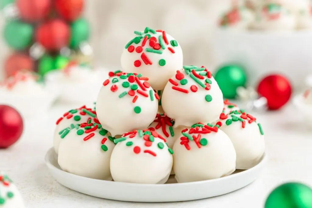 A plate of white sugar cookie truffles topped with festive red and green sprinkles, stacked in a pyramid shape. Holiday-themed decorations with blurred red and green ornaments are visible in the background.